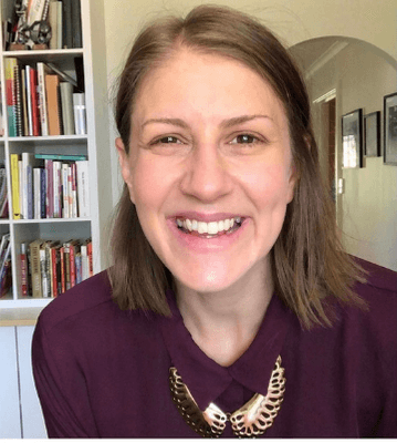 Smiling woman with light brown hair wearing a purple shirt and gold wing-shaped necklace.